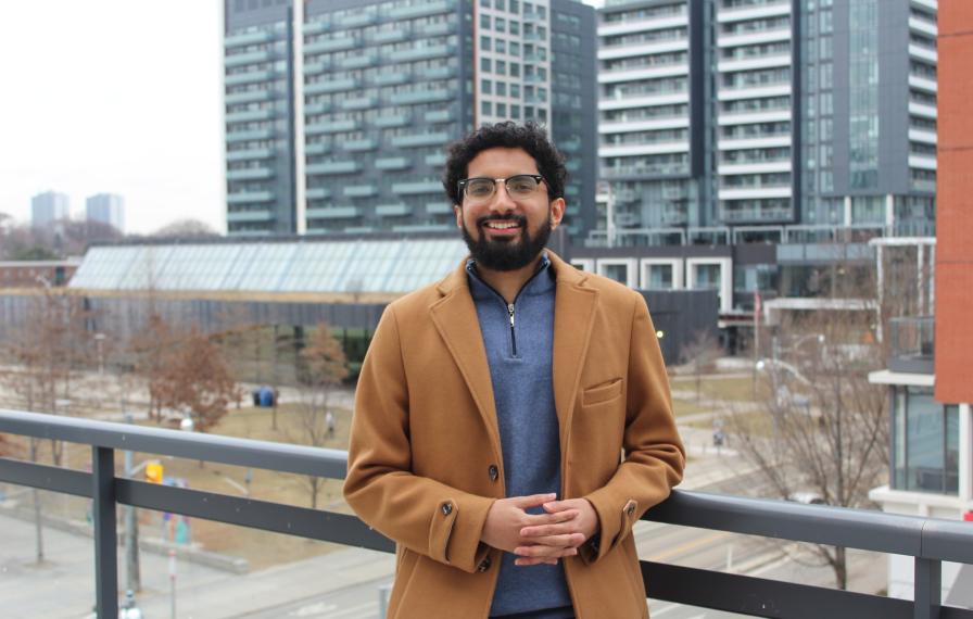 A person wearing an light brown coat and blue sweater standing on the balcony