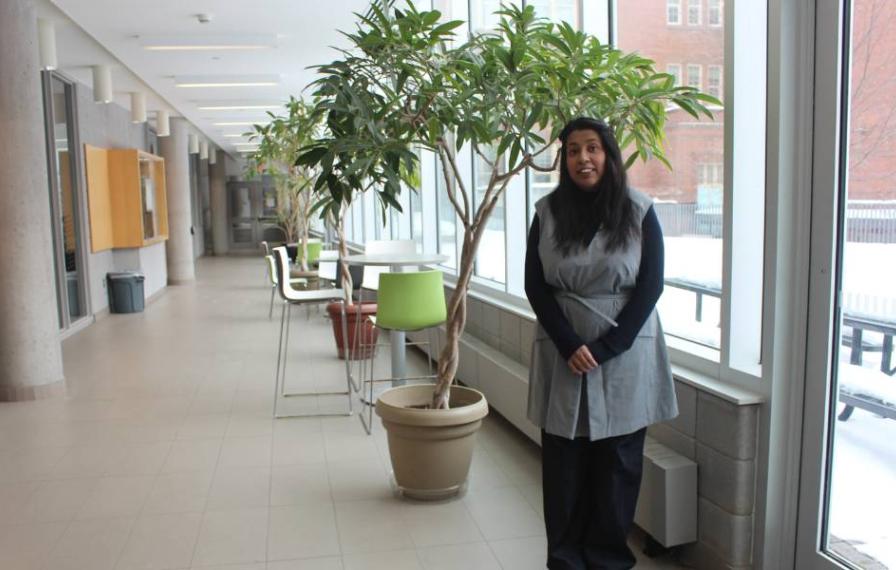 A woman smiling and standing in a hallway by windows and a small tree.