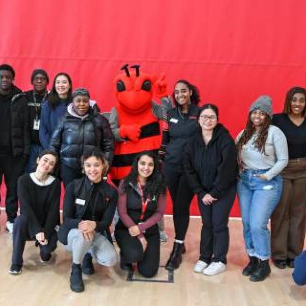 A group of students stand in front of a red wall. There is a red bee mascot in the middle.