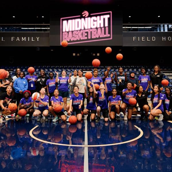 Team photo of the League of HER Own participants lined up on the basketball court, and throwing their basketballs towards the camera. In the background is a 'Midnight Basketball' sign.