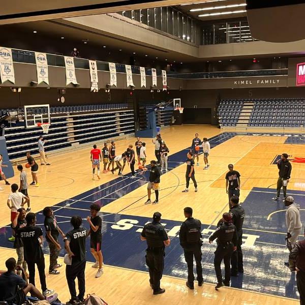 An indoor basketball court is filled with people. Some people are standing along the sidelines, chatting. Groups of young people are on the court playing informal games of basketball. A light sign that reads 'Midnight Basketball' can be seen across the court above the stands.