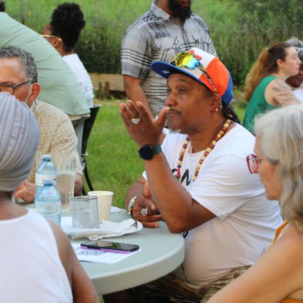 A man who is wearing a baseball cap is speaking to a group of people who are sitting at a table.