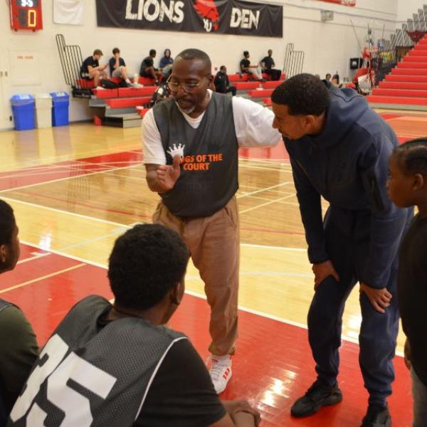 A person wearing a white shirt under a Black basketball jersey is speaking to a group of young men sitting on the bench. 