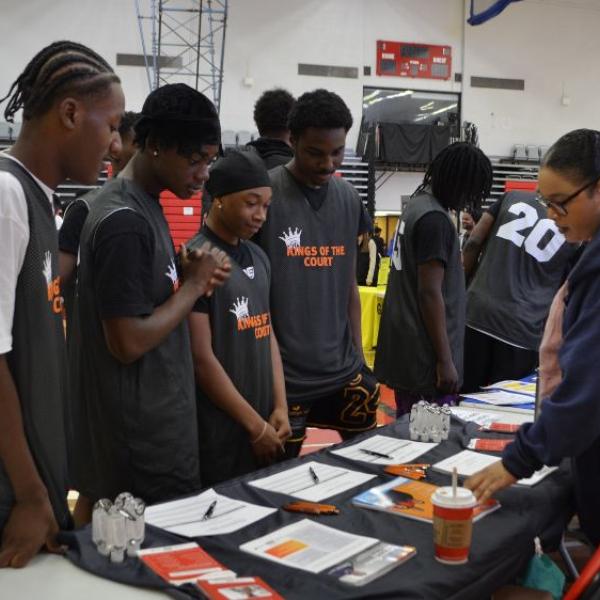 A group of boys wearing Black basketball jerseys is speaking to a woman wearing a purple sweater at the information table. 