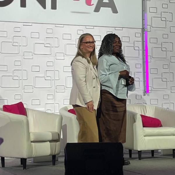 Two women on stage smiling at a camera. The woman on the right is holding an award plaque.