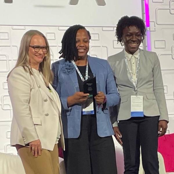 Three women standing on a stage smiling for a camera. The woman in the middle is holding an award plaque.