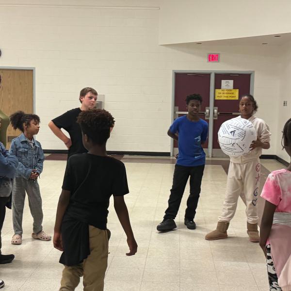 A group of individuals are standing in a circle. A girl, wearing a white sweatsuit, is holding a ball.   