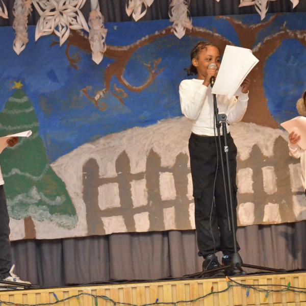 Three individuals holding scripts while speaking on stage.    