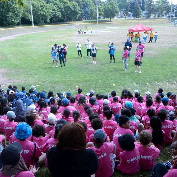 A group of children wearing blue and pink shirts sitting on a field listening to a presentation