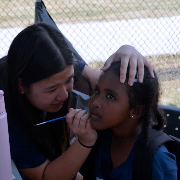 A young woman facing painting a child's face.
