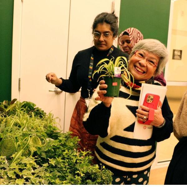A woman smiling and holding a small house plant, standing in front of a table with more house plants available to pick up.