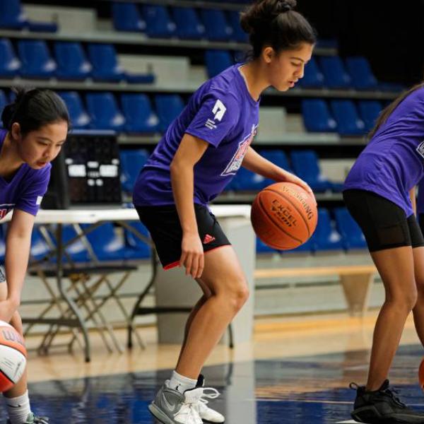 Girls standing on the side of a basketball court, practicing dribbling.