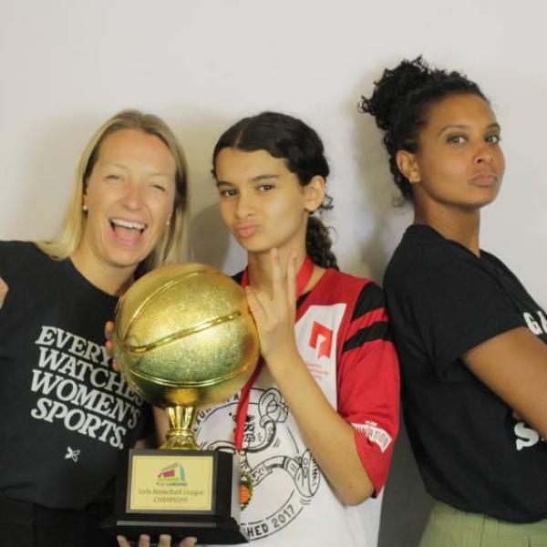 Three women holding a championship trophy and using their hands to show number 1.
