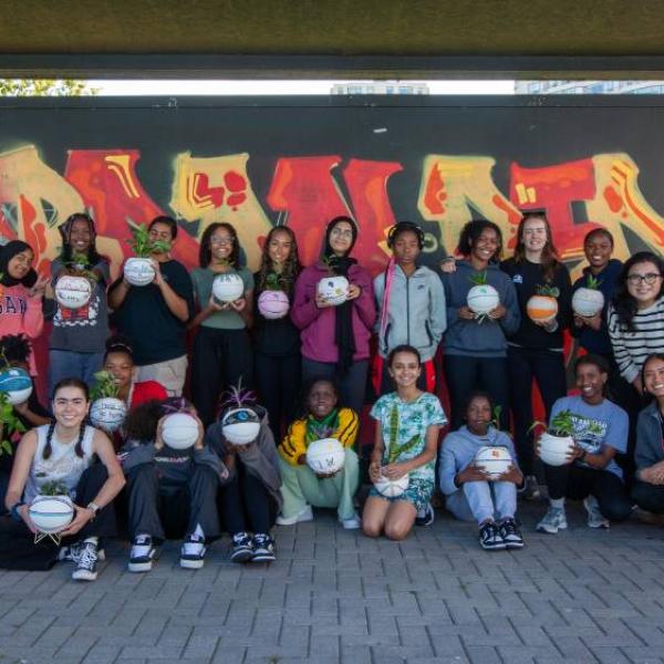 A large group of girls posing in front of a mural and holding painted basketball planters.