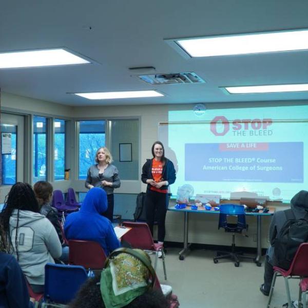 Two instructors present a "Stop the Bleed" course to an audience, with a projector displaying the course title. 