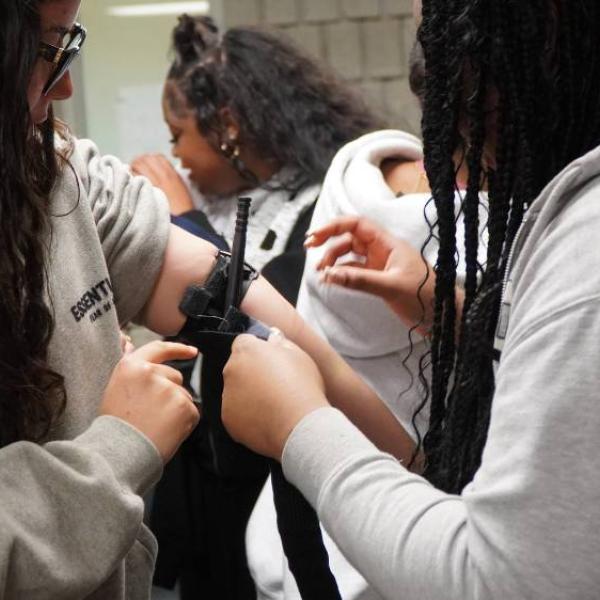 Two people gathered around as one person has a black tourniquet attached to their arm, while another person adjusts it.