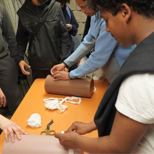 A close up of three people standing around a table. One person is giving instructions while the other two are packing wound with gauze. 