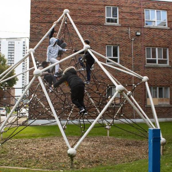 Children climb on a climbing structure in a park with an apartment building in the background.