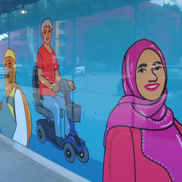 Close up of construction hoarding artwork showing a blue background with illustrations of people including a woman wearing a pink hijab, a person using an e-scooter, construction workers, and more.