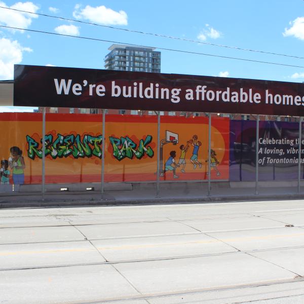 Construction hoarding artwork lining a street. The artwork features illustrations of people and the word "We're building affordable homes".