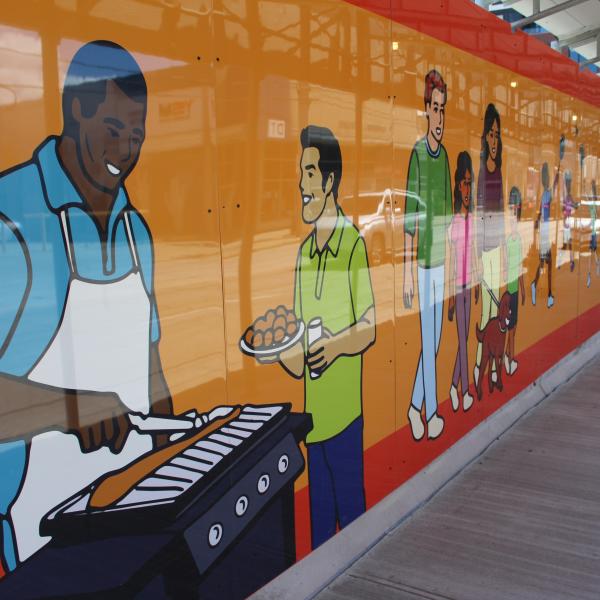 Brightly coloured construction hoarding showing illustrations of a man barbecuing, people and families walking and chatting on an orange background.