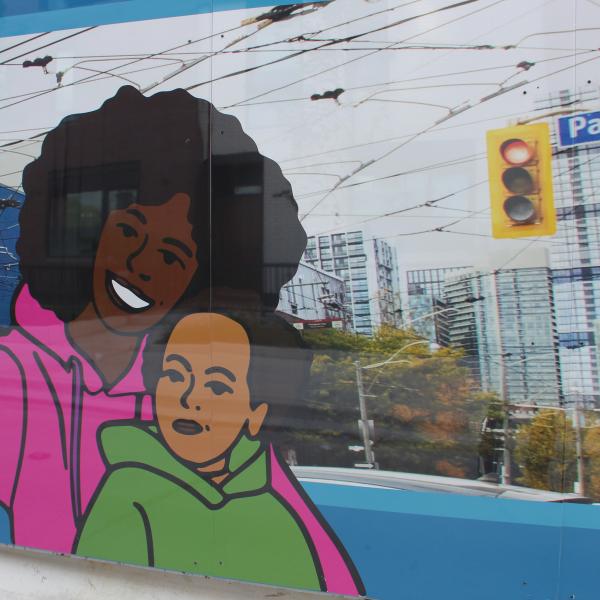 A close up of construction hoarding showing an illustration of a woman with a child taking a selfie overtop of a photography of the intersection at Parliament Street.