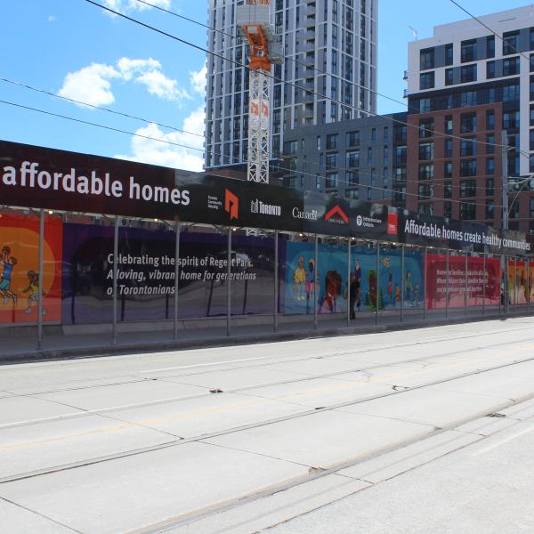A long view of construction hoarding along Gerrard Street showing brightly coloured artwork and buildings in the background.