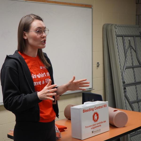 A woman in a red shirt and hoodie gives a lecture in a classroom. There is a first aid box on the desk.