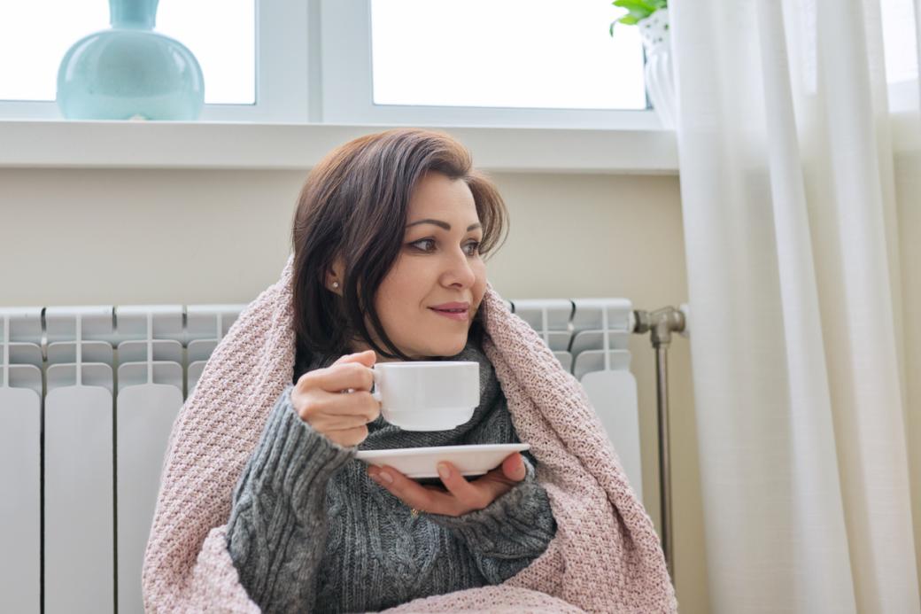 A woman warming up near home heating radiator while drinking tea.