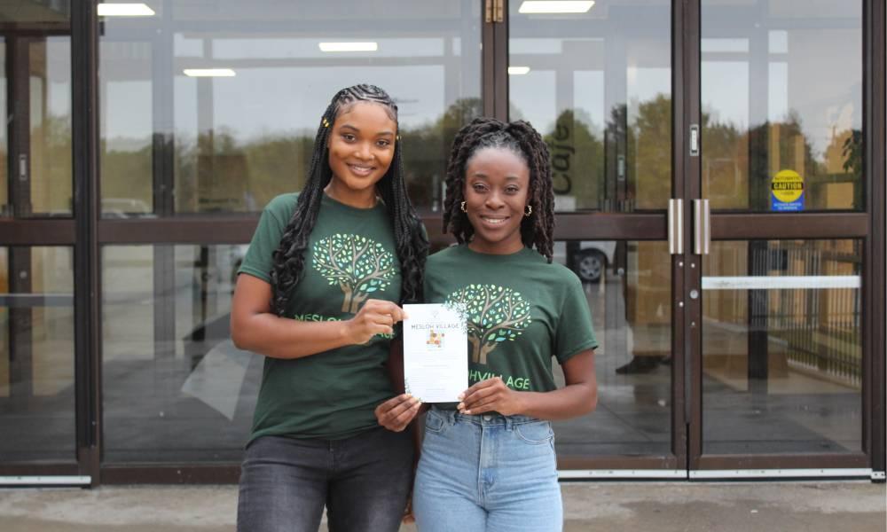 Sashalee Carty and Tamika Johnson standing outside the doors of a building holding a flyer about Mesloh Village.