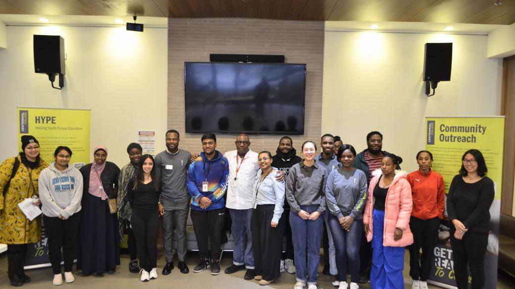 A group of diverse people smiling. They are standing in front of Centennial College banners. 