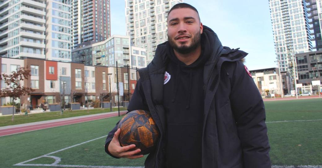 A man holding a soccer ball standing on a soccer pitch with high rise buildings in the background.