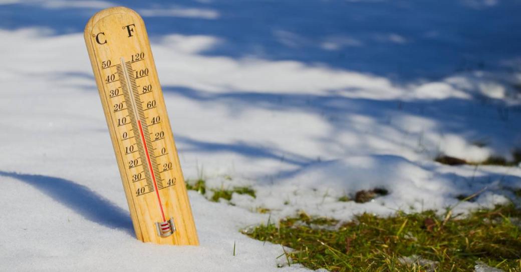 A thermometre sticking out of snow on the ground, while grass peeks through because the temperatures are hotter and the snow is melting.