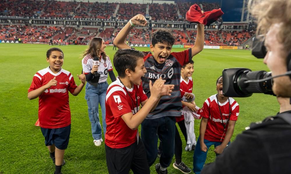 Five kids wearing TFC soccer jerseys running, jumping and smiling on BMO Field.