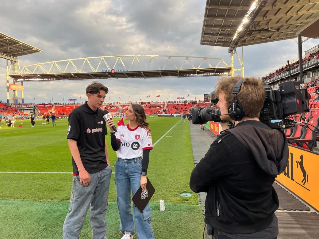 A woman interviewing a male on a soccer field. There is a videographer who is recording the interview.
