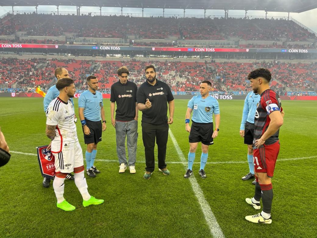 A group of men on the soccer field. A man wearing a Black shirt is conducting the coin toss.