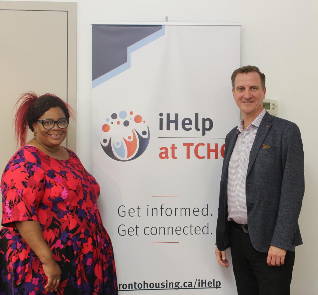 A Black woman in a red and blue floral dress poses with the CEO of TCHC, a white man, who is wearing a grey jacket and white shirt in front of the iHelp Centre banner. 