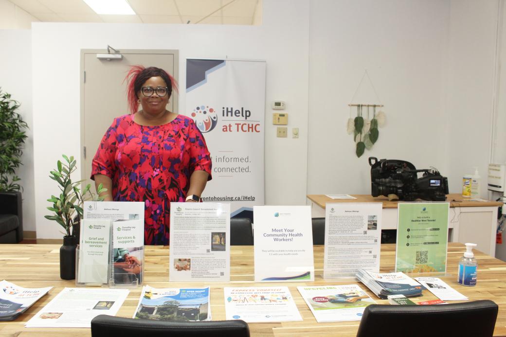 A Black woman in a red and blue-coloured floral dress stands behind a desk of iHelp Centre material. 