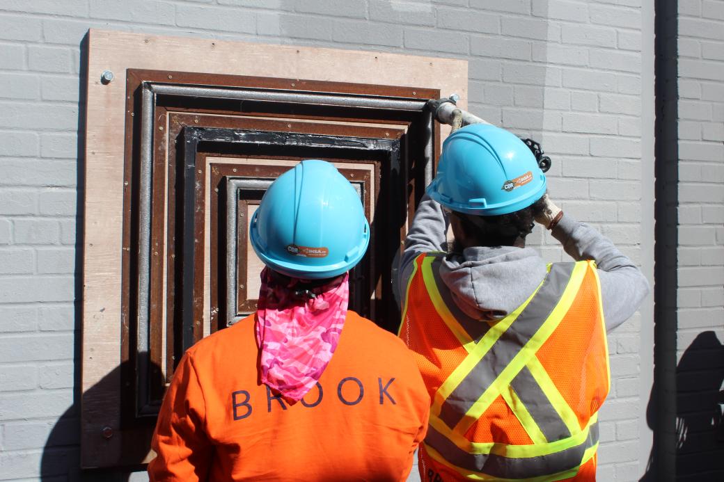 Two people wearing helmets are drilling into a window frame