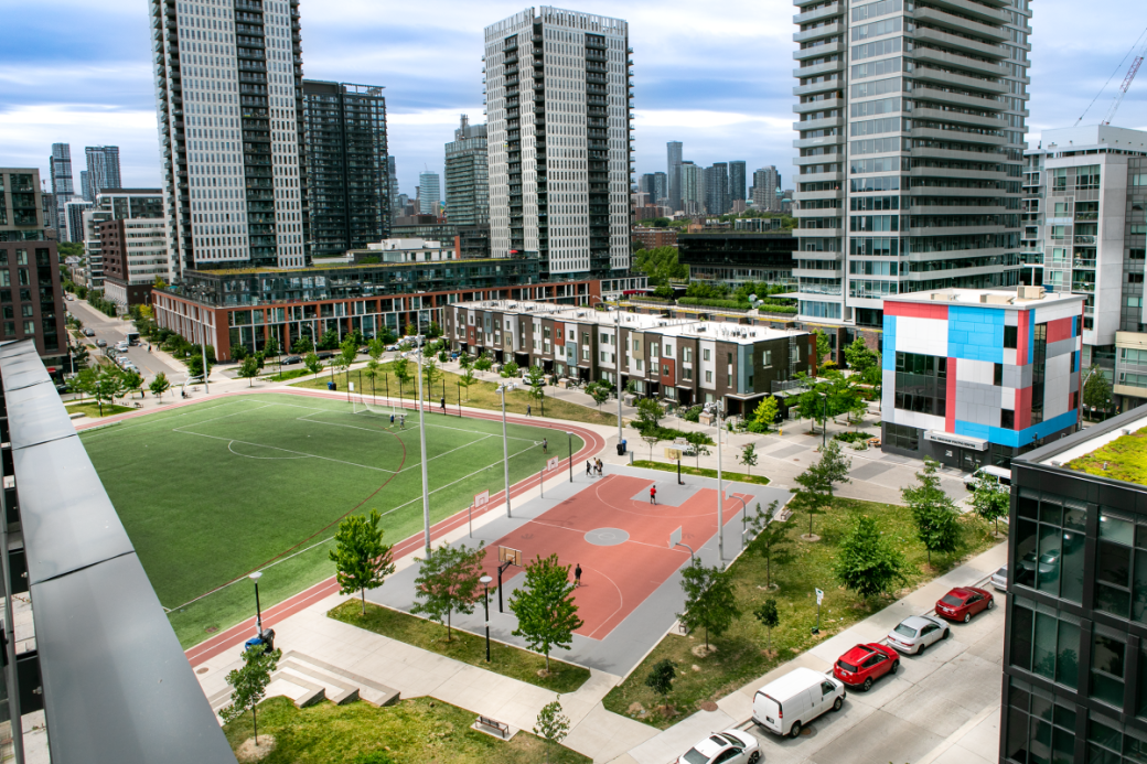 Photo credit: The Daniels Corporation An aerial view over Regent Park buildings, basketball court, and large field on a sunny day.