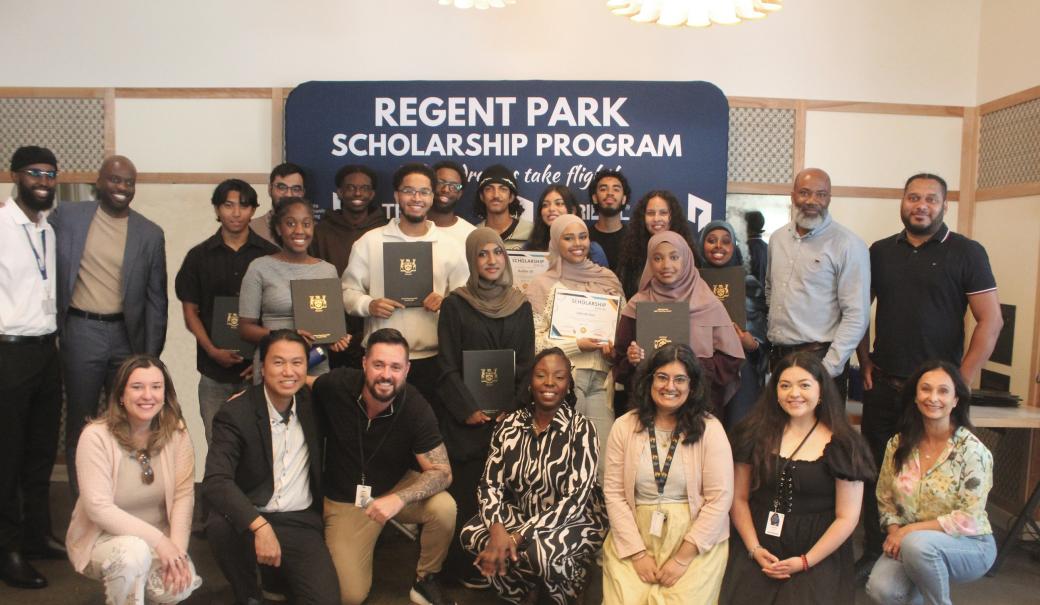 A group of people standing in front of the Regent Park Scholarship blue background and posing for a camera.
