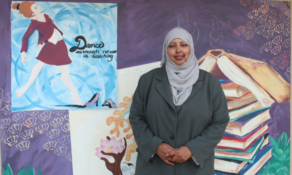 A woman standing in front of a mural showing a girl dancing with the words "Dance as though no one is watching"