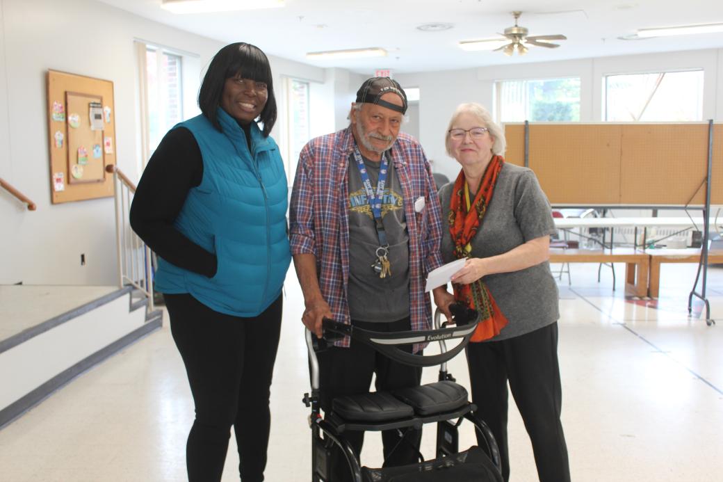 Mustafa in a mobiity device is flanked by two TCHC staff posing - the staff member on the left is handing Mustafa a white envelope. 