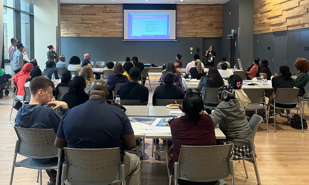 A large group of people sitting at tables and chairs in a community room, looking at a screen with a presentation.
