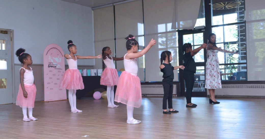 A group of young Black girls wearing pink ballet outfits and tutus practicing ballet..