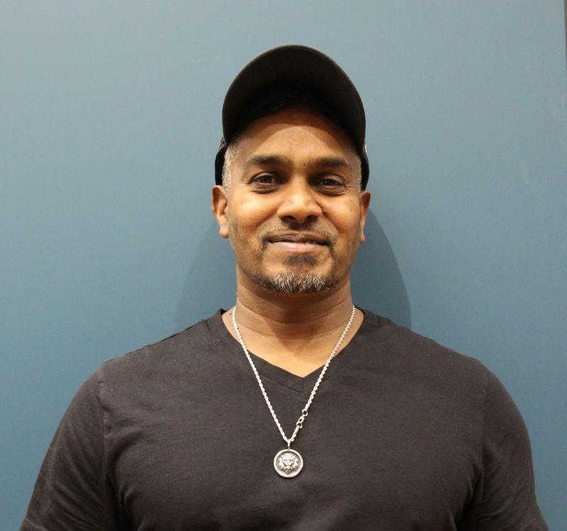 A man wearing a baseball hat smiling, standing against a blue wall.