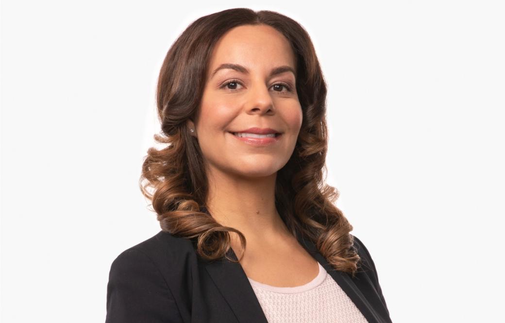 A photo of a woman with long brown hair wearing a black blazer standing in front of a white background smiling.