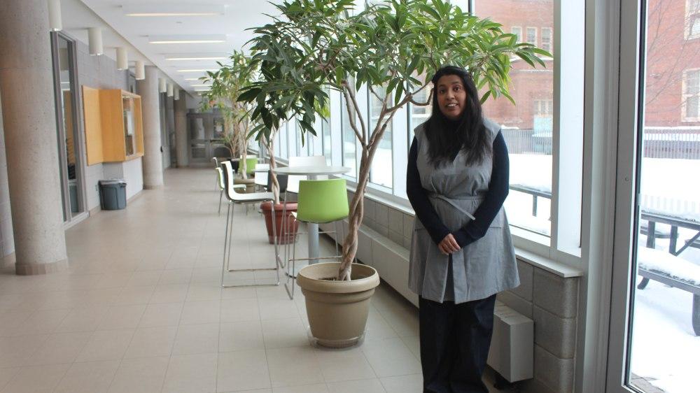 A woman wearing a grey dress stands in a hallway with roundtables, chairs and plants in the background.