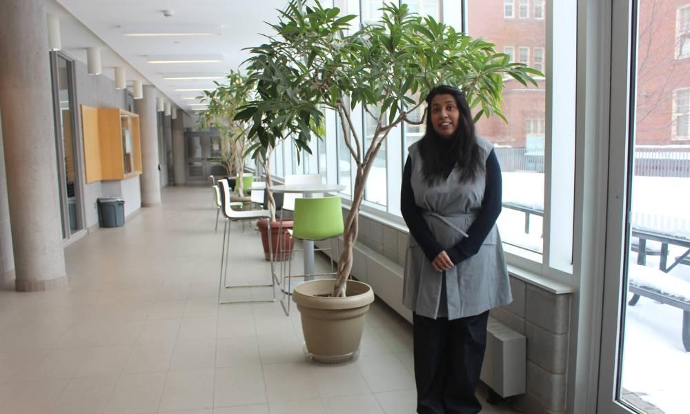 A woman smiling and standing in a hallway by windows and a small tree.