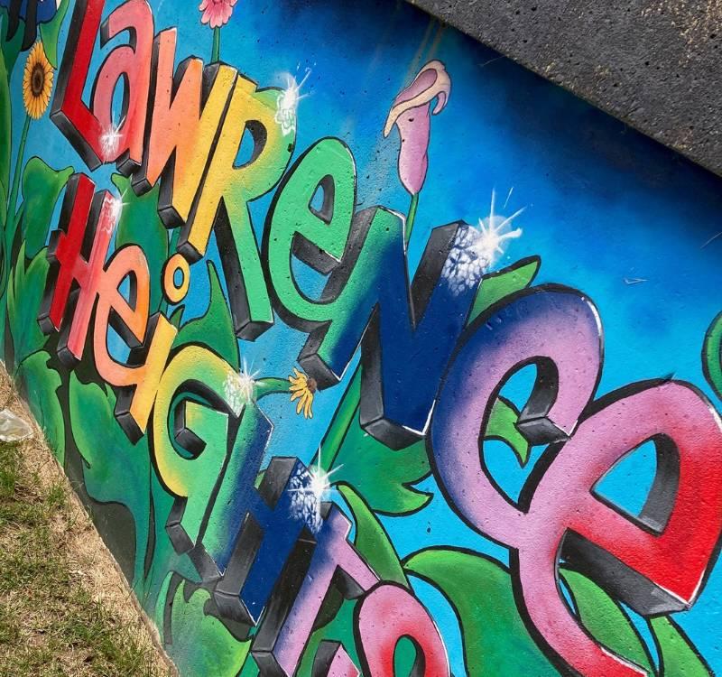 A section of a mural with the words 'Lawrence Heights' painted in rainbow colours. The words appear on top of green plant stems and leaves, with a blue sky in the background.